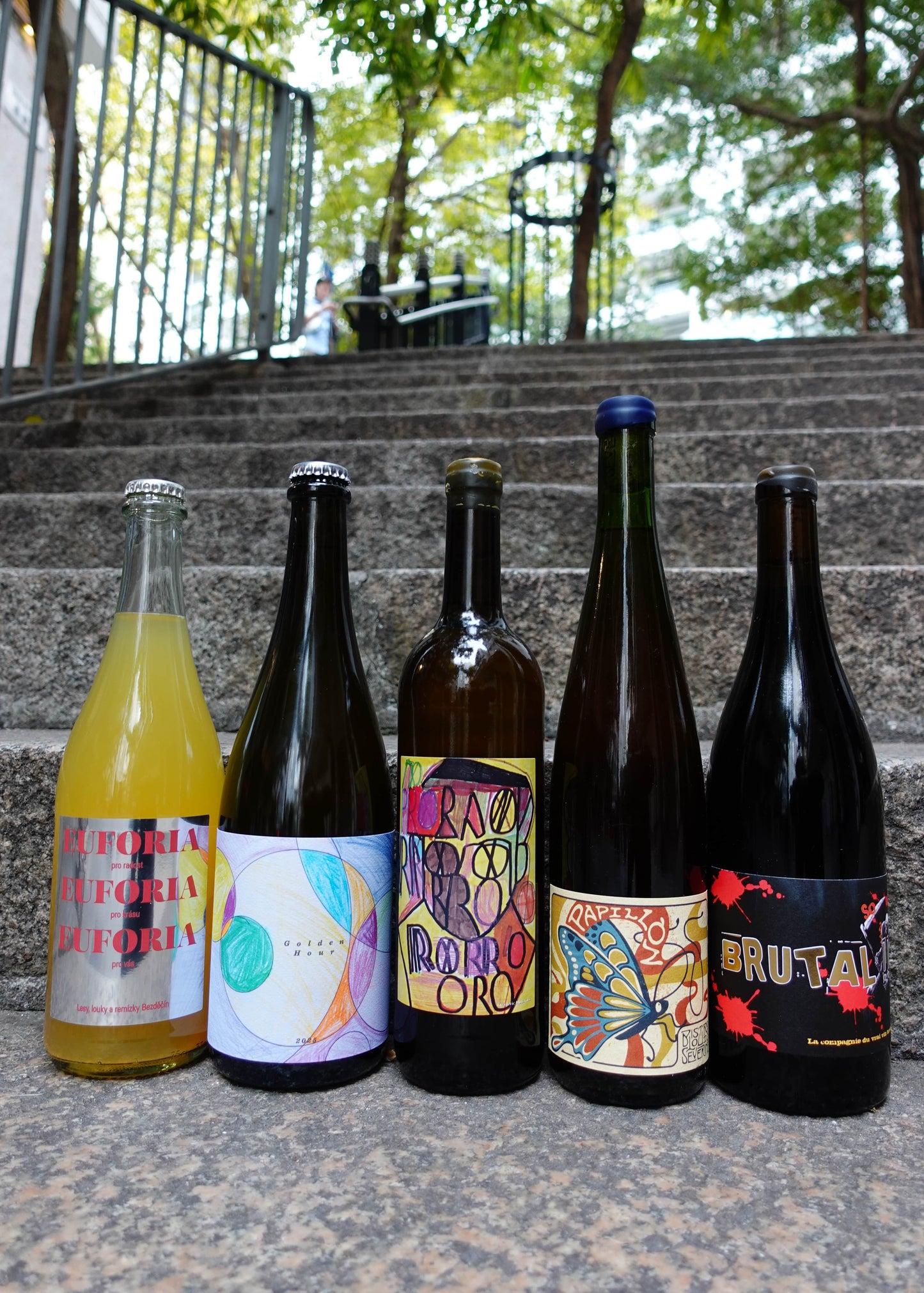 Five bottles of different types of drinks on a set of outdoor stairs with a blurred background.